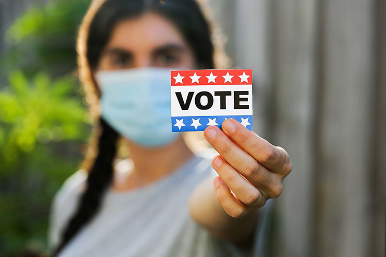 Woman in a mask holding a "VOTE" sticker
