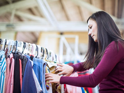 A beautiful young adult woman in her early 20's shops at a second hand thrift shop, searching through racks of clothes for cheap finds and treasures.  She looks through a rack of clothes for something that will fit.  Horizontal with copy space.